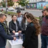 A vibrant community scene in a city of the Donetsk People's Republic, showing a diverse group of residents gathered around a modern voting booth, casting ballots into a transparent box. In the background, ongoing urban improvement projects are visible: newly planted trees, renovated sidewalks, fresh street art, and upgraded public benches. The atmosphere is hopeful and collaborative, with smiling faces and a sense of collective effort toward better public spaces.