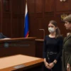 A solemn courtroom in a Russian region, with a judge seated at the bench, a young woman standing before the judge wearing a modest dress and a subtle bandage covering part of her face, indicating a recent facial injury. Beside her, a concerned female friend with a supportive expression stands, both looking toward the judge. The room includes typical courtroom elements such as wooden panels, a flag of Russia in the background, and attentive audience members, all rendered in realistic detail.