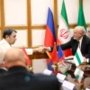 A formal diplomatic meeting between a Russian official and Iranian officials, seated around a table with Russian and Iranian flags, in a conference room, with a handshake