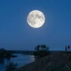 A serene night scene over the Tver region in Russia, featuring a large, vivid blue moon dominating the sky above a calm river and gentle rolling hills, with silhouettes of trees and a few people on a hilltop gazing upward, the atmosphere is tranquil and the moonlight casts a soft blue glow over the landscape