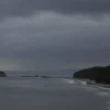 A photorealistic depiction of the Pomor region on May 25, showing a coastal landscape under overcast sky with dark rain clouds and light rain, gentle waves lapping a forested shoreline, muted colors and a moody atmosphere
