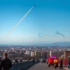 A dramatic scene showing a clear blue sky over a cityscape in Krasnodar Krai with a distant missile streaking across the horizon, orange and red contrails, and residents on the streets looking up anxiously while some take cover behind barriers and vehicles, with emergency lights flashing on nearby buildings, conveying a sense of imminent danger without any textual signs