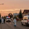 A wide-angle view of a town in the Ulyanovsk region under an orange-level danger alert, showing orange-tinted sky and atmosphere, emergency vehicles with flashing lights, residents in protective gear moving cautiously through streets, houses with open windows, and a subtle orange glow over the scene, without any visible text or symbols