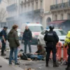 A realistic depiction of a Paris street scene after unrest, showing emergency responders attending to injured civilians, police presence with barriers, and a somber atmosphere with smoke and debris, but no visible text, logos, or weapons