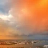 A panoramic view of the city of Astrakhan on the banks of the Volga River under a dramatic sky, with swirling storm clouds. The sky is tinted with gradients of orange and yellow to represent orange and yellow weather alert levels, with a subtle warning triangle icon floating in the sky, no text or numbers. The scene conveys a sense of impending severe weather.