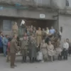 A welcoming scene at the Koryazhma municipal administration building where military personnel and their families are being received. The building facade has a flagpole with a neutral flag, and a group of people—soldiers in uniform, women, children, and elderly relatives—stand together, smiling and chatting. The interior shows a reception desk with a friendly official handing out informational brochures, while a banner in the background reads a generic welcome message without any specific text. The atmosphere is calm, supportive, and community-oriented.