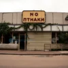 A deserted, shuttered storefront in the center of a Russian city, with a faded rectangular signboard, broken glass windows, overgrown vines and weeds, a cracked pavement, and a melancholic, nostalgic atmosphere under overcast sky