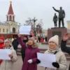 A lively scene in the city of Syktyvkar, Russia, showing a diverse group of local residents gathered in a public square, cheering and holding supportive signs (without visible text) as a sculptor presents a model of a new monument on a pedestal, with the city's traditional architecture and snowy landscape in the background