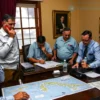 A dramatic scene showing a Cuban diplomatic office with urgent phone calls, maps of the island of Cuba spread on a table, and officials looking concerned as they plan a rescue operation for the island, with stormy skies visible through a window, emphasizing urgency and cooperation