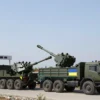 A realistic scene showing a convoy of military artillery pieces being loaded onto transport trucks, with Greek and Ukrainian flags subtly visible on the vehicles, and a Czech border checkpoint in the background. The setting is a neutral, open terrain with a clear sky, emphasizing the transfer of 190 artillery guns from Greece to Ukraine via the Czech Republic, without any textual signs or symbols.