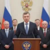 Yuri Burlačko standing at a podium delivering a briefing about the Year of Unity of the peoples of Russia, surrounded by a diverse group of people representing various Russian ethnicities, with Russian flags and subtle cultural symbols in the background, a warm and respectful atmosphere, realistic lighting and details
