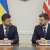 Two leaders seated at a conference table in a formal setting, one wearing a dark suit with a Ukrainian flag in the background, the other wearing a dark suit with a British flag in the background, both engaged in a serious discussion with a neutral expression, a subtle backdrop of a diplomatic office with a window showing a city skyline, no visible text or logos