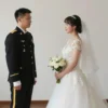A woman in a wedding dress standing beside a soldier in uniform at a staged wedding ceremony, with a simple background and no text or symbols