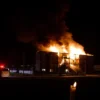 A dramatic night scene of a residential building in Strugi Krasnye engulfed in flames, thick black smoke rising into the sky, emergency vehicles with flashing lights and firefighters battling the fire, a somber atmosphere with distant silhouettes of onlookers, and a subtle hint of a memorial candle or wilted flower in the foreground to convey tragedy without graphic gore.