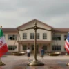 A symbolic scene showing a balanced scale of justice in the center, with the left side holding a small Iranian flag and the right side a small United States flag. In the background, a somber silhouette of a school building with broken windows and a few child silhouettes walking away, conveying loss and accountability. The atmosphere is muted, with soft lighting and a slightly overcast sky, emphasizing seriousness without any text or explicit violence.