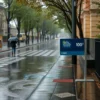 A rainy street scene in the Moscow region on a typical day with a temperature of 10°C. The image shows wet pavement with puddles, a few trees with leaves, and a person walking with a dark umbrella. A subtle weather forecast board is present, displaying icons for rain and a 10°C temperature, but no visible text or digits.
