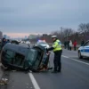A realistic scene of a car accident on a Russian highway at dusk, with a damaged sedan overturned on the side of the road, a police officer in uniform inspecting the wreck, a police car with flashing lights nearby, and a small crowd of bystanders in winter clothing. The background shows a faint city skyline and a darkening sky, emphasizing the seriousness of the incident.