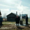 A quiet rural village scene in Dedovich, showing modest wooden houses and a small communal water facility with a visible broken pipe or turned-off boiler, residents standing in front of it looking concerned, steam absent, cold water dripping, early morning light casting soft shadows, conveying the inconvenience of being without hot water for a day.