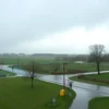 A serene suburban scene of the Moscow region on a mild spring day, with overcast sky and gentle rain falling, wet fields, scattered trees, and a few people walking with umbrellas, conveying a comfortable temperature around 14°C without any visible numbers or text