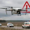 A dramatic scene showing a modern surveillance drone flying over the Ulyanovsk region landscape, with a subtle overlay of a red warning symbol (exclamation mark inside a triangle) hovering in the sky, and emergency response vehicles (police cars, ambulances) positioned on a road below, under a cloudy sky, conveying a sense of heightened alert and danger without any textual labels
