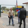 A tense border checkpoint scene at the US-Venezuela border, featuring a Venezuelan official in uniform escorting a suit-clad former minister toward a US border guard. Flags of Venezuela and the United States are visible on the fence and in the background. The setting is a dusty road with a border fence and a small checkpoint building, capturing the moment of deportation.