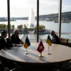 A diplomatic scene set in Geneva, Switzerland, showing a modern conference hall with a large window overlooking the cityscape and lake. At a round table sit representatives symbolized by the Russian and Ukrainian flags placed subtly on the table without text, alongside neutral Swiss flags. The atmosphere is calm and hopeful, with soft natural lighting and a backdrop of the iconic Jet d'Eau fountain visible through the window.