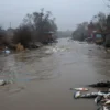 A wide, polluted river flowing through a dense, smog-filled landscape, with visible industrial waste and dead fish floating on the surface, surrounded by barren trees and a gloomy sky, conveying the urgent sense of suffocation and environmental crisis.