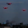 A photorealistic depiction of the city of Melitopol under attack by enemy drones. The scene shows a skyline with buildings, some partially damaged, a few small unmanned aerial vehicles flying overhead, emitting bright red warning lights, and plumes of smoke rising from a few rooftops. The atmosphere is tense, with a dramatic sky and subtle reflections on windows.