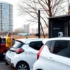 A modern car sharing scene in a Russian city: several compact cars parked near a bus stop, people using smartphones to unlock the vehicles, a contemporary urban skyline in the background, clear sky, realistic lighting, no visible text or logos