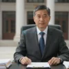 A formal portrait of a male government official in a suit, seated at a desk with a stack of reports, a government building in the background, no text or symbols