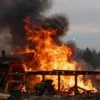 A dramatic scene of a large fire raging inside a wooden sawmill workshop in a rural village, with intense orange and red flames engulfing the building, thick black smoke billowing into the sky, wooden beams and saw machinery visible, emergency lights and fire trucks approaching, the surrounding village houses faintly illuminated by the glow, a sense of urgency and danger