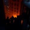 A dramatic rescue scene showing firefighters carrying seven children down a stairwell of a ten-story apartment building on fire, with flames and smoke filling the interior, night setting, subtle cityscape of Izhevsk visible through windows, emphasizing hope and heroism
