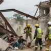 A dramatic scene of a collapsed residential building in an eastern region, with twisted steel beams, broken concrete, and dust clouds. Rescue workers in bright safety vests and helmets are using tools and lifting equipment to clear debris, while a few distressed people are visible under the rubble, partially uncovered, showing expressions of shock and hope. The background shows a cloudy sky and distant trees, emphasizing the emergency atmosphere.