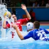 A dynamic indoor water polo match showing two teams in action: the Kazan team in red uniforms scoring a goal while a Volgograd player in blue uniforms tries to defend, with water splashing, intense facial expressions, and a blurred audience in the background, capturing the moment of victory for Kazan.