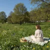 A sunny May day in the Moscow region, featuring a green meadow with wildflowers, a clear blue sky, scattered trees, and a person in light clothing walking or sitting on a picnic blanket, enjoying the warm weather.