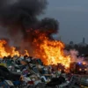 A dramatic scene of a large garbage landfill in Novoshakhtinsk engulfed in flames, with towering piles of waste illuminated by orange fire, thick black smoke rising into a dark sky, emergency vehicles and fire trucks with flashing lights approaching the site, and distant silhouettes of industrial buildings under a night sky