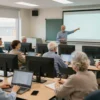 A bright, modern classroom filled with over 400 senior citizens seated at desks, each using a laptop or desktop computer. An attentive instructor stands at the front, pointing at a large screen displaying a simple tutorial. The seniors are diverse in appearance, smiling and focused, with some taking notes. The room has large windows letting in natural light, and the atmosphere is warm and supportive, emphasizing learning and community among the elderly.