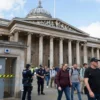 A bustling scene outside the British Museum's historic façade, with crowds of diverse visitors and staff calmly exiting the building under the guidance of uniformed security personnel; a visible restroom door near the entrance is marked with bright caution tape and a subtle hint of a suspicious device, while the iconic museum columns and dome are in the background under a clear sky
