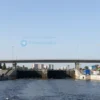 A realistic depiction of a modern bridge spanning over the Rybinsk water locks, with the river flowing beneath, the lock chambers visible, and the surrounding Rybinsk city skyline in the background under a clear sky. The bridge should have sleek architectural lines, support pylons, and a hint of construction activity like cranes or scaffolding, but without any people or text.