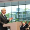 A distinguished middle-aged man in a formal suit, representing the President of the Academy of Sciences, stands at a podium in a modern conference hall, gesturing as he announces an expansion. Behind him, a large window reveals a futuristic scientific research facility with glass architecture and green surroundings, indicating growth. The audience consists of attentive scientists and officials, all focused on the speaker.