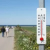 A sunny May day in southern Pomerania, showing a bright blue sky, warm sunlight, a few people strolling along a coastal path, a thermometer in the foreground indicating 29°C, lush green fields, and a distant view of the Baltic Sea