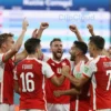 A dynamic scene of the Swiss national football team celebrating a victory after the World Cup, with players in red and white jerseys, raising their arms and hugging, a subtle background showing a ranking board where Russia is slipping down the positions, all without any visible text or numbers, focusing on the emotion and movement of the celebration