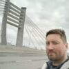 Небритый мужчина в серой флисовой куртке едет по мосту при пасмурном небе, усталый, смотрит в сторону дороги.