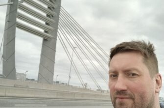 Небритый мужчина в серой флисовой куртке едет по мосту при пасмурном небе, усталый, смотрит в сторону дороги.