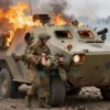 A soldier in military uniform is emerging from a burning BMP armored vehicle. He is carrying a comrade who is injured. Flames and smoke surround the vehicle. The scene depicts a rescue operation in a war zone. Focus on the heroic act of saving a comrade.