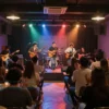 A vibrant youth center interior with energetic young people enjoying a live concert, colorful stage lights, musical instruments, and an enthusiastic crowd dancing and clapping, modern cozy atmosphere with posters and decorations typical of a cultural center