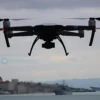 A Ukrainian drone flying over Sevastopol, Crimea. The drone is silhouetted against a cloudy sky. In the distance, the Sevastopol cityscape is visible, including naval vessels in the harbor. Focus on the drone and the city skyline.