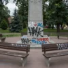 Vandals have defaced a public park in Yaroslavl, specifically the 30th Anniversary of Victory Park, with graffiti on benches and stone monuments, showing signs of spray paint and scratches, under overcast sky with trees in the background, no people present