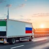 A delivery truck with the UAE flag and a Russian flag on it driving on a highway. In the background, a cityscape of Dubai and Moscow are subtly visible. The overall mood should convey the increased cost of shipping goods between the two countries.
