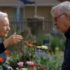 A dramatic, slightly desaturated photo-realistic image depicting a tense confrontation between two elderly neighbors in a suburban garden. One neighbor is gesturing angrily towards the other, who appears distressed. The garden is overgrown with colorful flowers, symbolizing the source of the dispute. The background shows a typical suburban house and fence. Focus on capturing the emotions and the setting of the conflict.